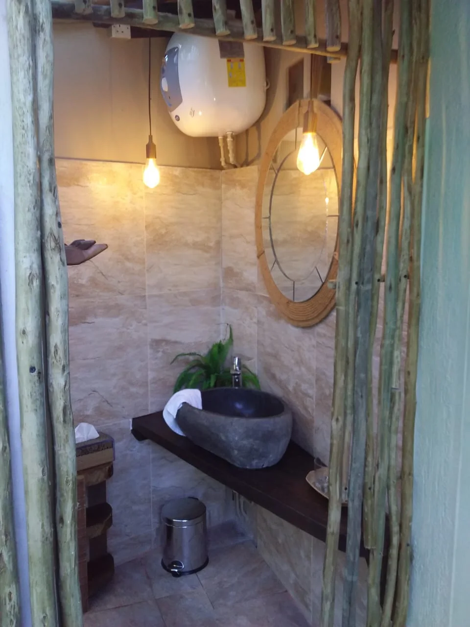 Rustic ensuite bathroom with stone vessel sink, wooden pole doorway and circular mirror with wooden slats — Garden Suite 2 at Maison Soleil, Anse Soleil, Seychelles