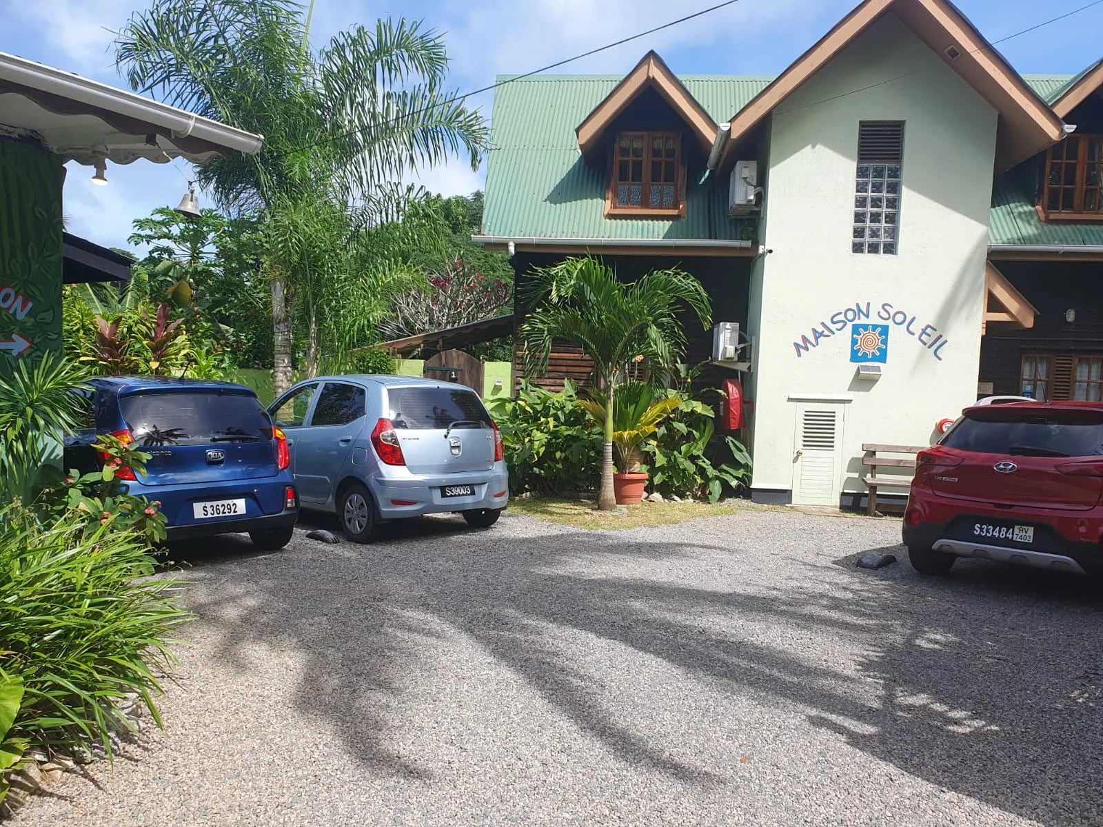 Maison Soleil main building with sun logo, green roof, wooden wings and guest parking — boutique guest house facade at Maison Soleil, Anse Soleil, Seychelles